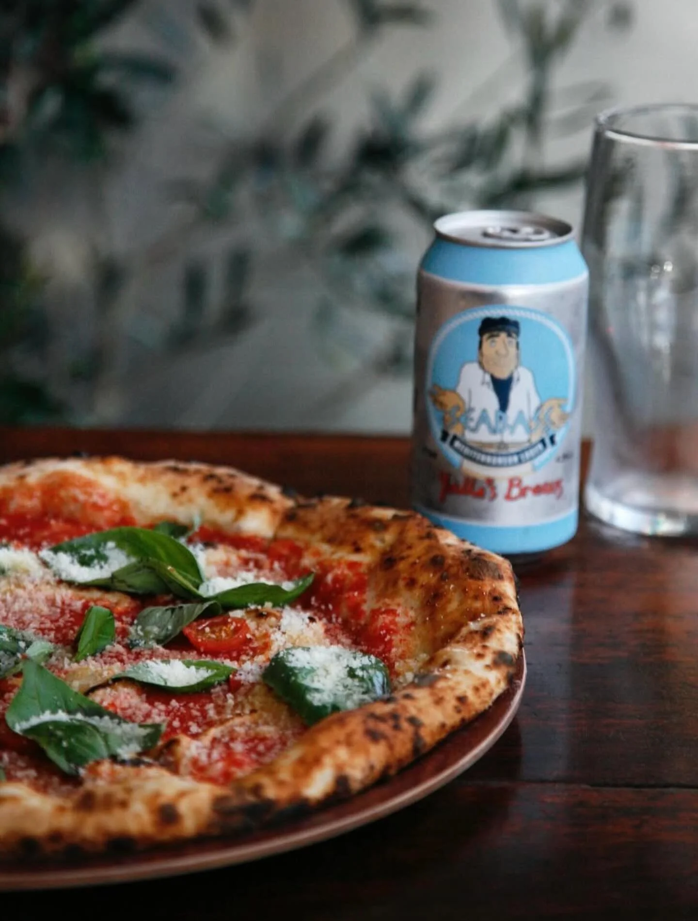 A pizza with tomato sauce, fresh basil leaves, and grated cheese on a wooden table, with a can of soda and a glass in the background.