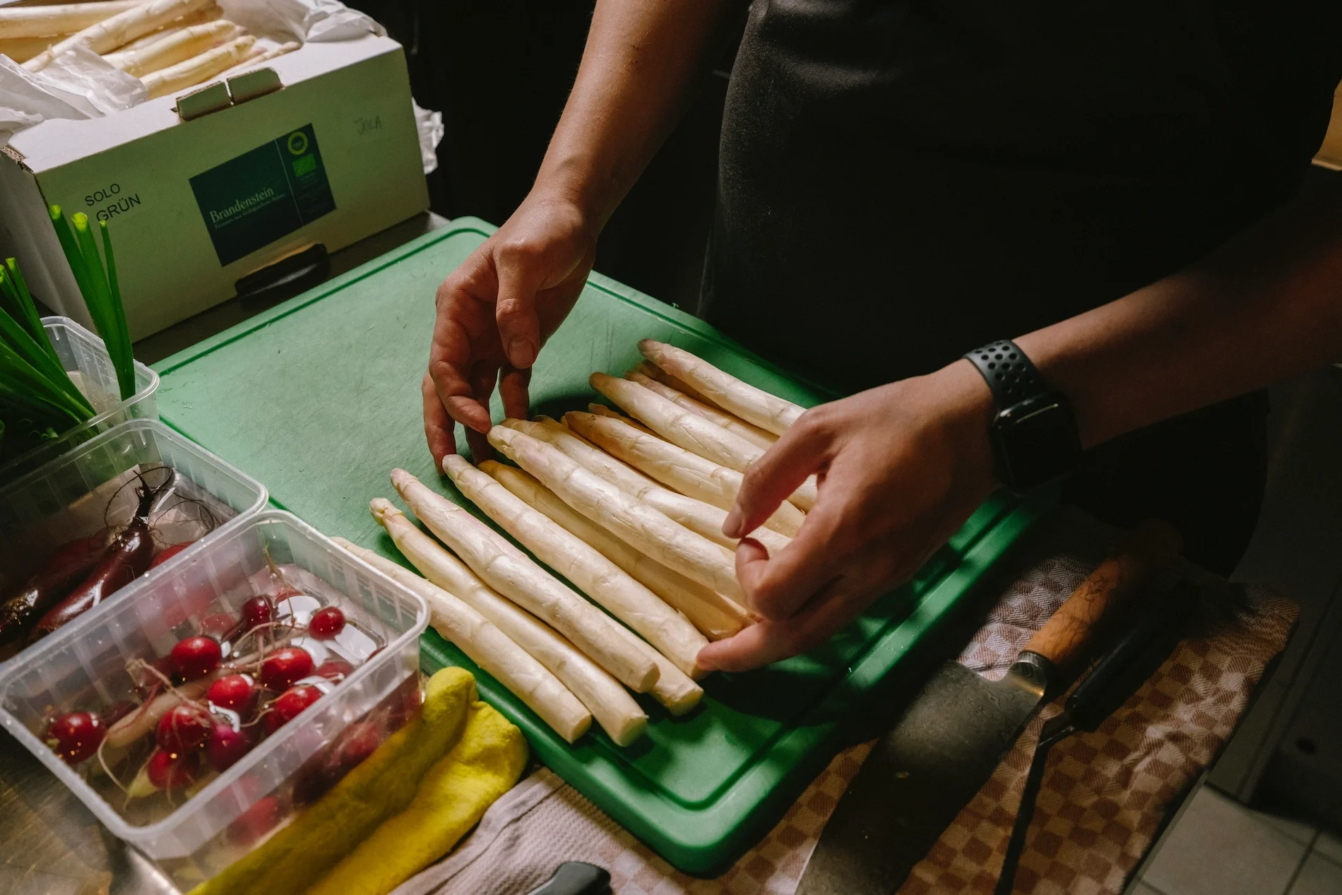 A person preparing white asparagus spears on a green cutting board, with cherries, yellow squash, and green onions visible in plastic containers nearby.
