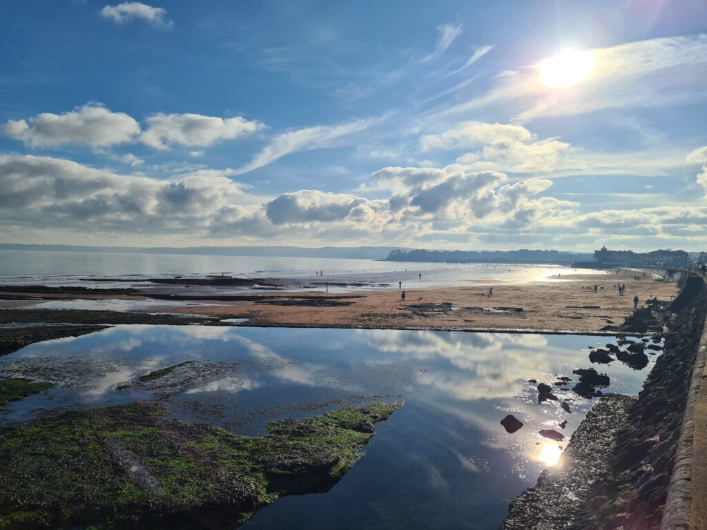 A beach scene with a partly cloudy sky and a setting sun, people walking along the shoreline, and calm water reflecting the sky and clouds.