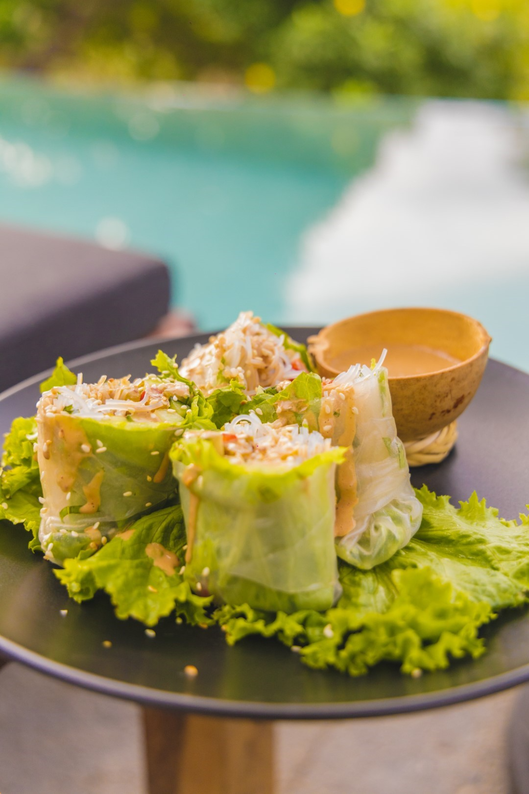 Fresh spring rolls with lettuce, rice noodles, and shredded chicken on a black plate, garnished with sesame seeds, with a small bowl of dipping sauce, poolside background.