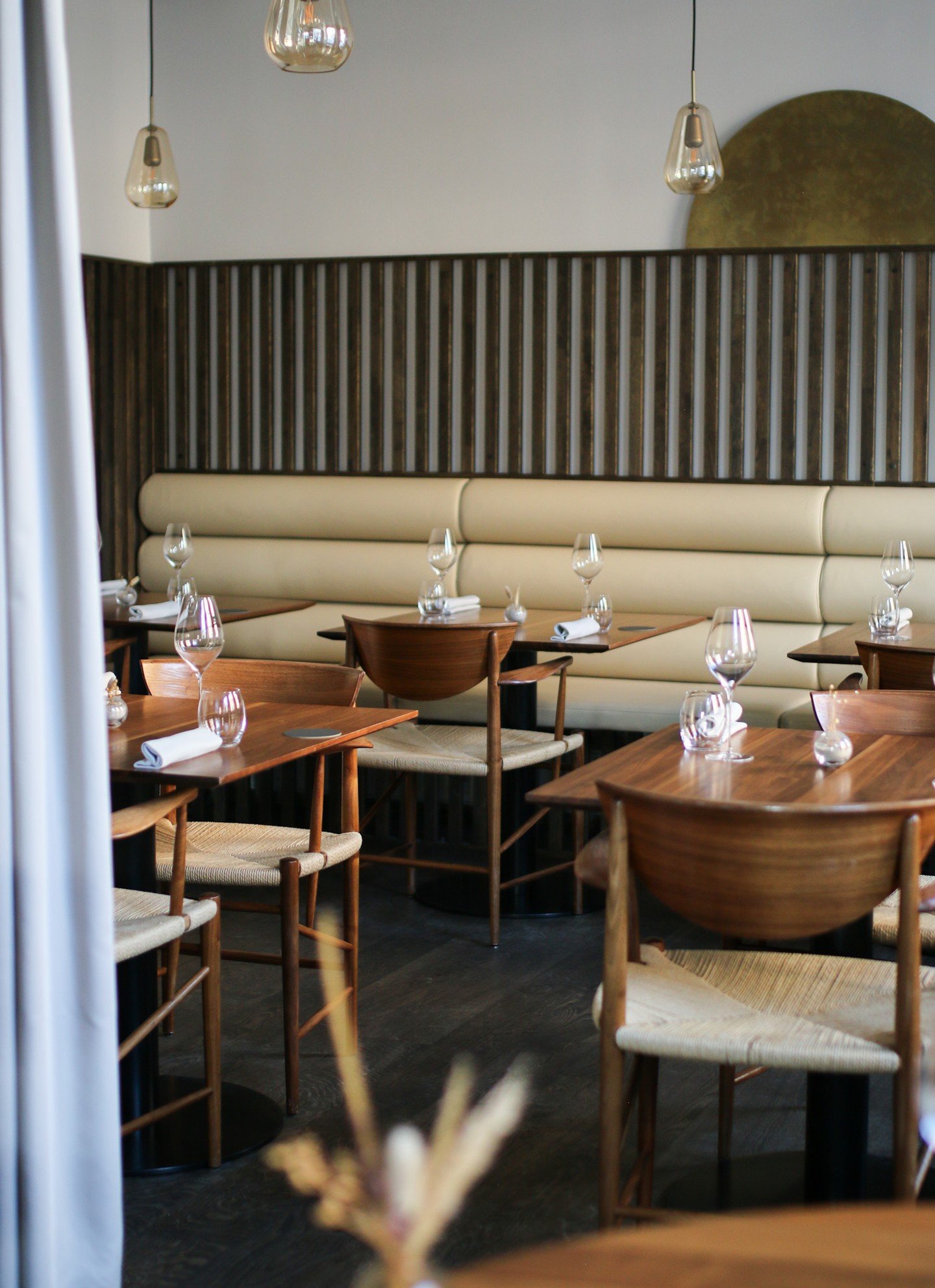 Interior of a modern restaurant with wooden tables, beige chairs, wine glasses, white napkins, a beige cushioned bench, pendant lights, a dark wooden slat wall, and a decorative wall feature.