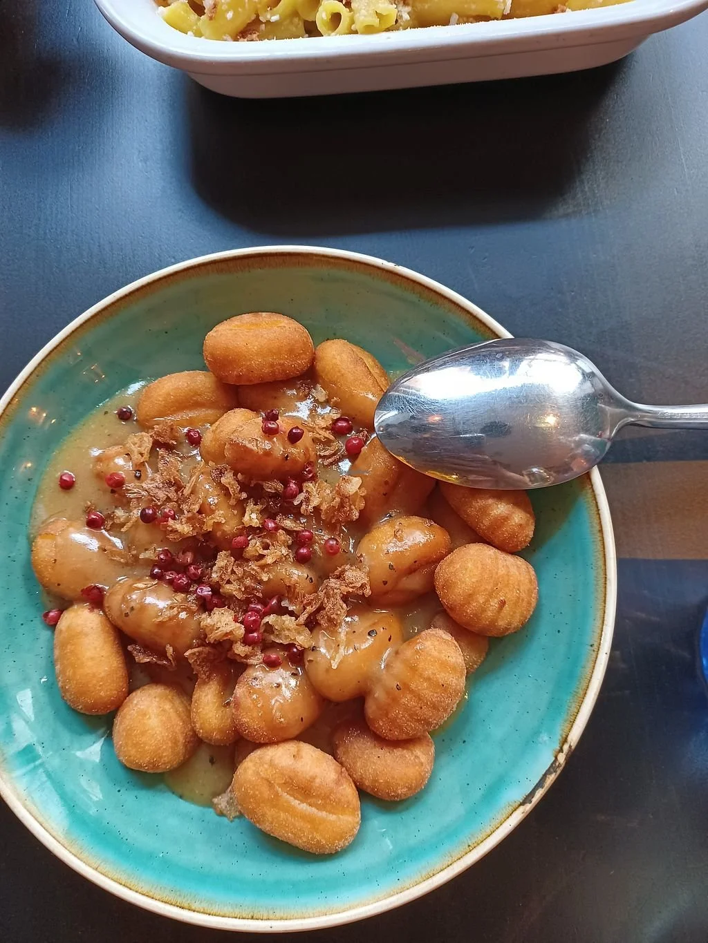 A bowl of breaded fried potato nuggets topped with gravy, pink peppercorns, and crispy onion bits, with a spoon resting on the side.