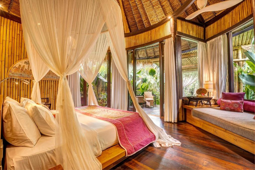 A tropical bedroom with bamboo walls, a four-poster bed with white curtains and bedding, and a sitting area with cushions and a sofa, surrounded by lush greenery outside.