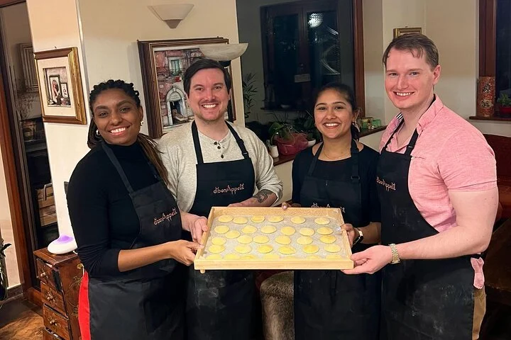 Four people wearing aprons standing together in a home kitchen, holding a tray of uncooked round dough pieces, smiling at the camera.