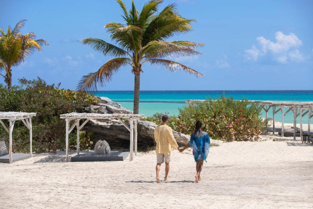 A couple holding hands walking on a sandy beach with palm trees, greenery, and cabanas, facing the ocean with a clear blue sky.