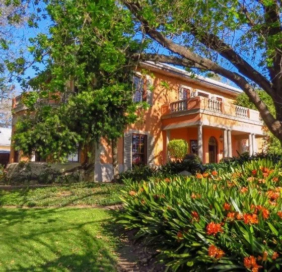 A large two-story house with a balcony and arched entrance, surrounded by green trees, a lawn, and flowering plants.