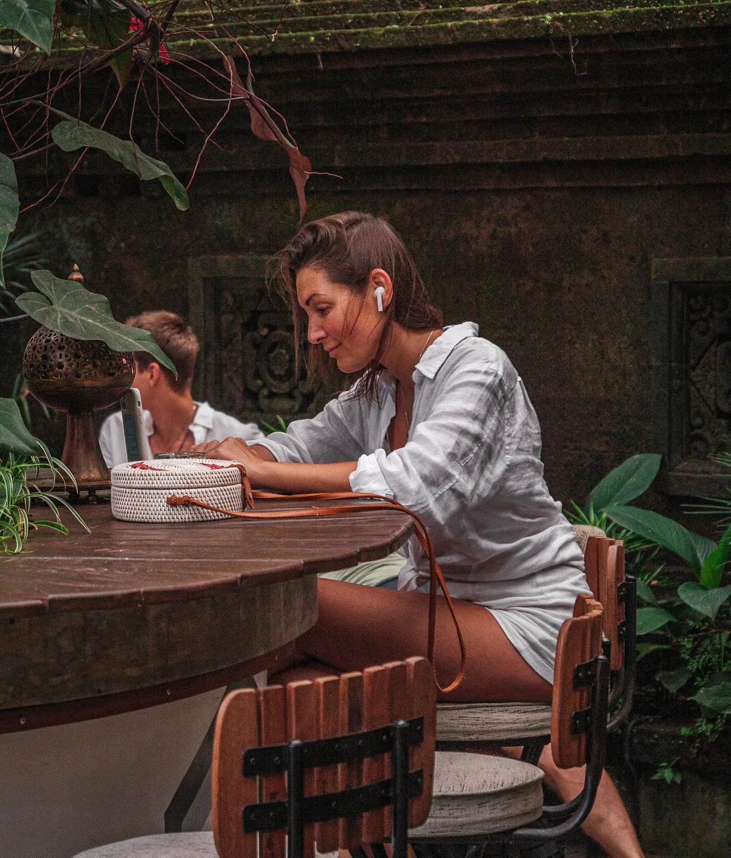 A woman with long brown hair wearing a white shirt and wireless earbuds, sitting at a wooden table in a lush, green setting, looking down at a round woven purse in front of her, with a man partially visible in the background.