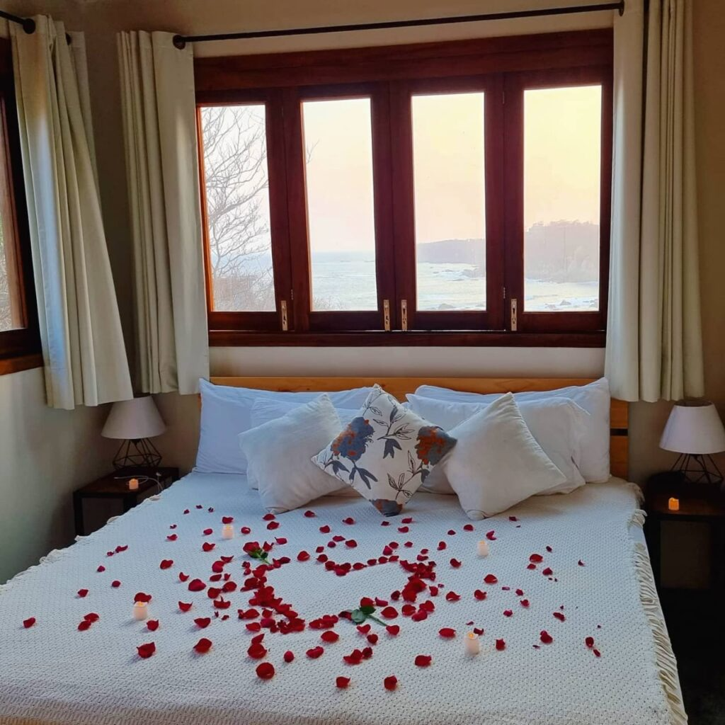 A cozy bedroom with a bed decorated with red rose petals forming a heart shape and small candles, overlooking a scenic view of water and hills through a large window.