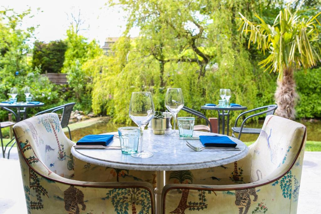 Outdoor dining table with two chairs, set for two with wine glasses, water glasses, and blue napkins. Background features lush green trees, bushes, and a small pond.