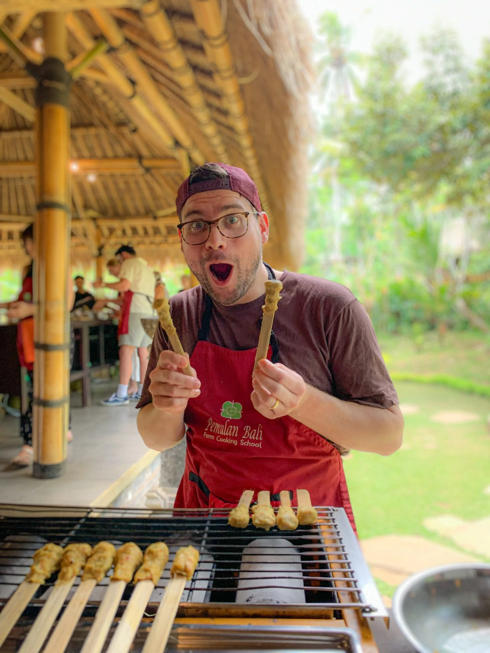 A man wearing glasses, a maroon cap, and a red apron is smiling with his mouth open while holding skewers of cooked food over a grill. There are other people in the background at an outdoor bamboo structure surrounded by green trees.