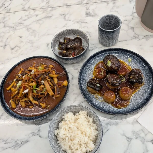 A meal featuring a bowl of white rice, a plate of braised beef with vegetables and sauce, a small bowl of cooked eggplant with sauce, and a plate of braised meatballs in sauce, all on a marble table.
