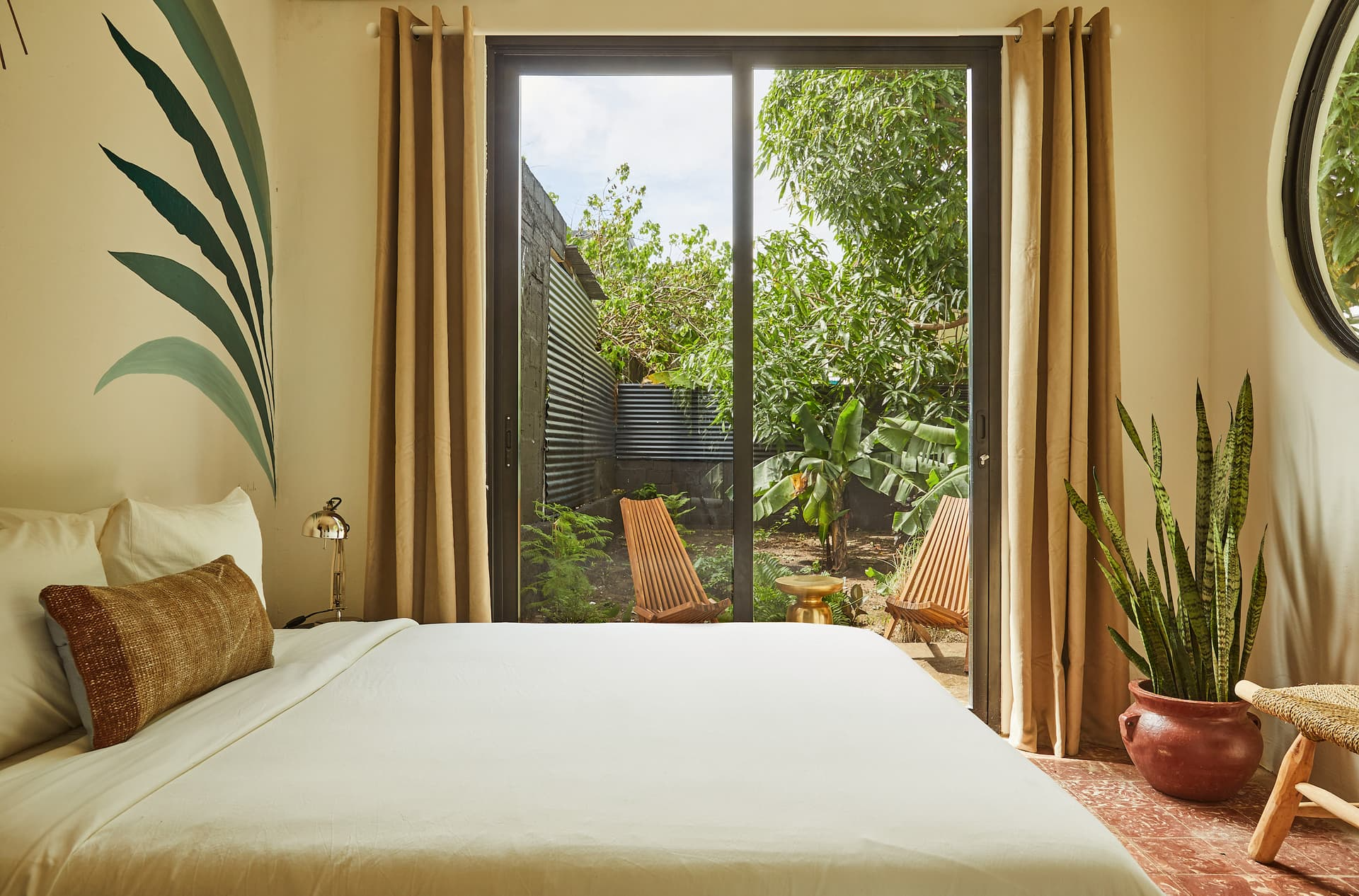 A bedroom with a sliding glass door leading to a garden with two wooden chairs, green plants, and trees outside. The room has a bed with a white comforter, a brown textured pillow, a small nightstand with a metallic lamp, and a potted plant on the right.