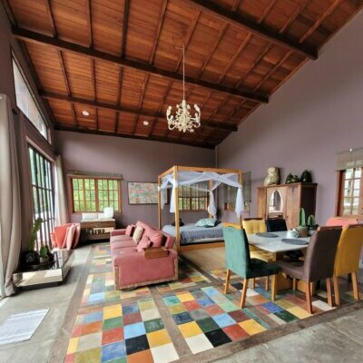 Bright living room with a colorful patchwork rug, pink sofa, four chairs around a dining table, and a canopy bed in the background, with large windows and wooden ceiling