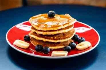 Stack of four pancakes topped with syrup and blueberries, served on a red plate with banana slices.