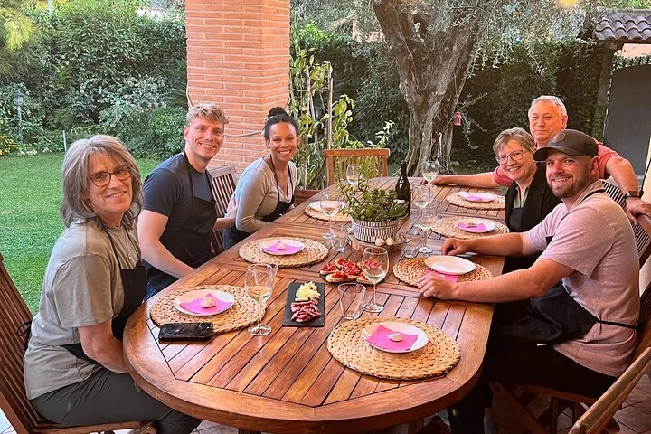 A group of seven people sitting around a wooden outdoor dining table, smiling, with plates, glasses, and a centerpiece