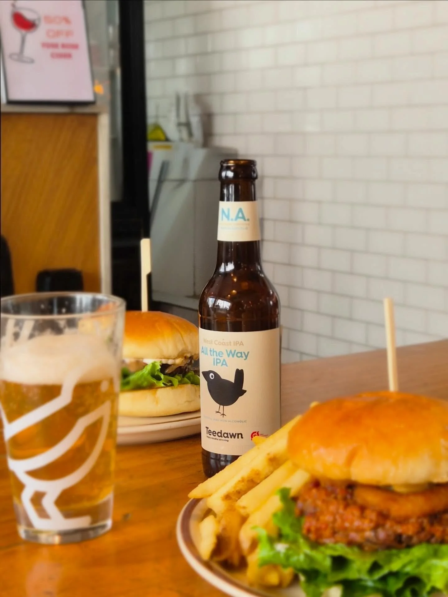 A table with a beer, a burger with lettuce, tomato, and a meat patty, a flatbread or sandwich, and a bottle of N.A. beer labeled 'All the Way IPA' in a restaurant with white tiled walls.