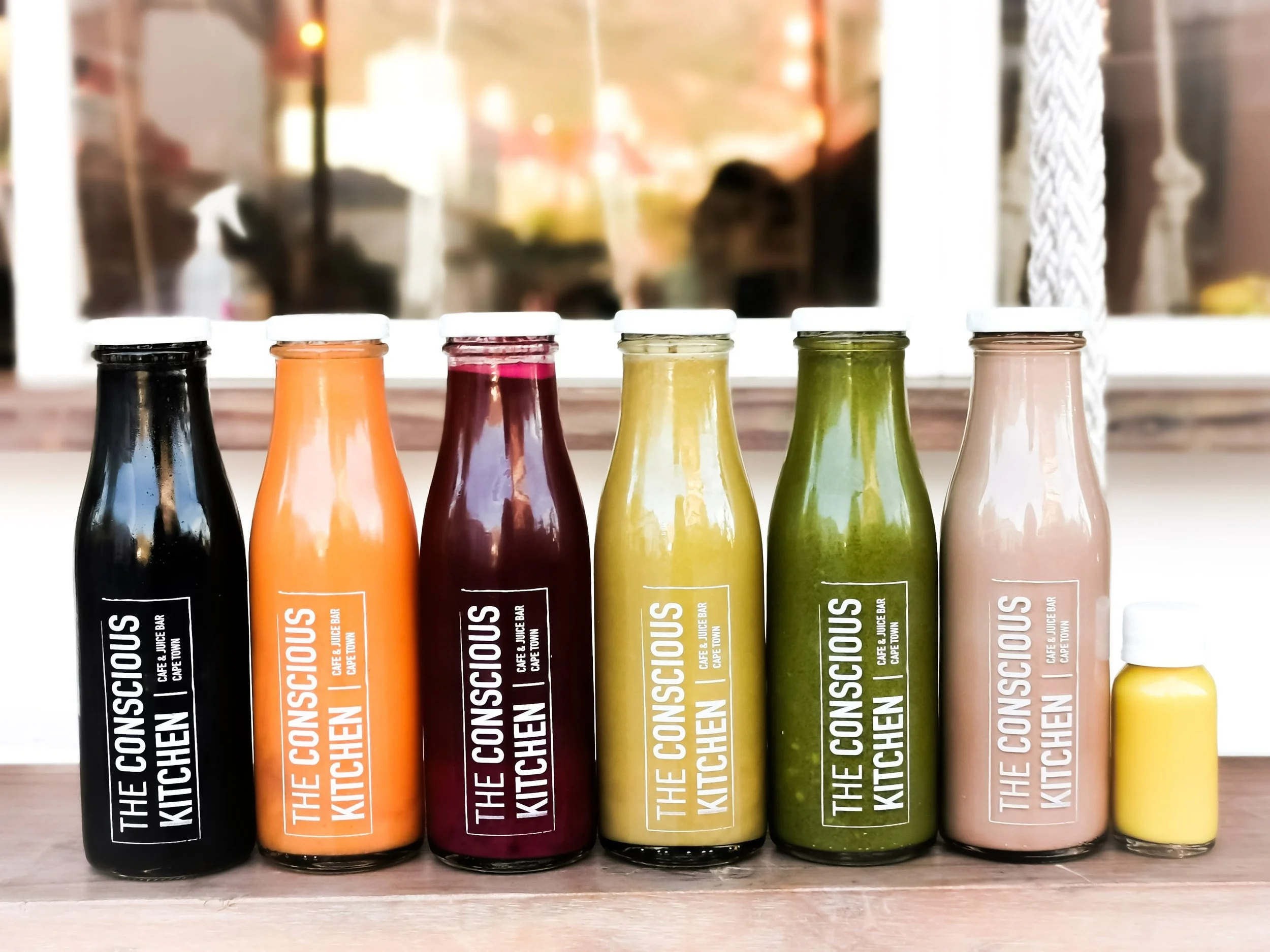A row of six bottles of colorful juice drinks with white caps, labeled with the brand name 'THE CONNOISSEUR KITCHEN' and different fruit or vegetable flavors, placed on a wooden surface with a blurry background.