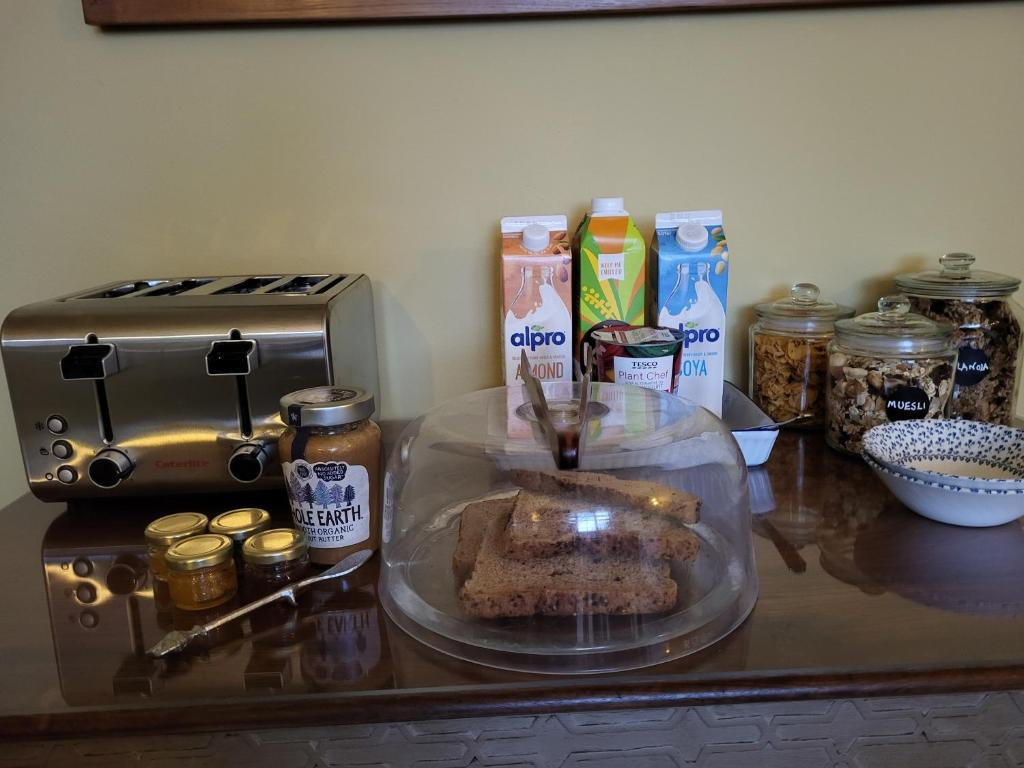 Breakfast setup on a wooden table with toaster, jars of jam, sliced bread under a glass cover, cartons of almond and soy milk, muesli in glass jars, a bowl of cereal, and a small container of plant-based spread.