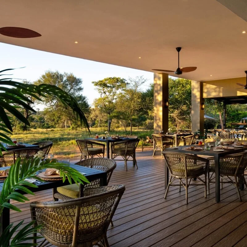 Outdoor patio dining area with tables and chairs, overlooking a natural landscape with trees and greenery, under an overhanging roof.