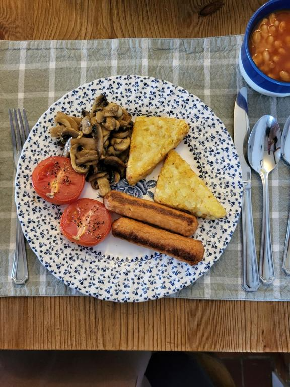 A plate with fried eggs, sautéed mushrooms, sliced tomatoes, potato waffles, and vegetarian sausage links. A side of baked beans in a bowl is on the upper right. The meal is set on a checkered placemat with a fork, knife, and spoon.