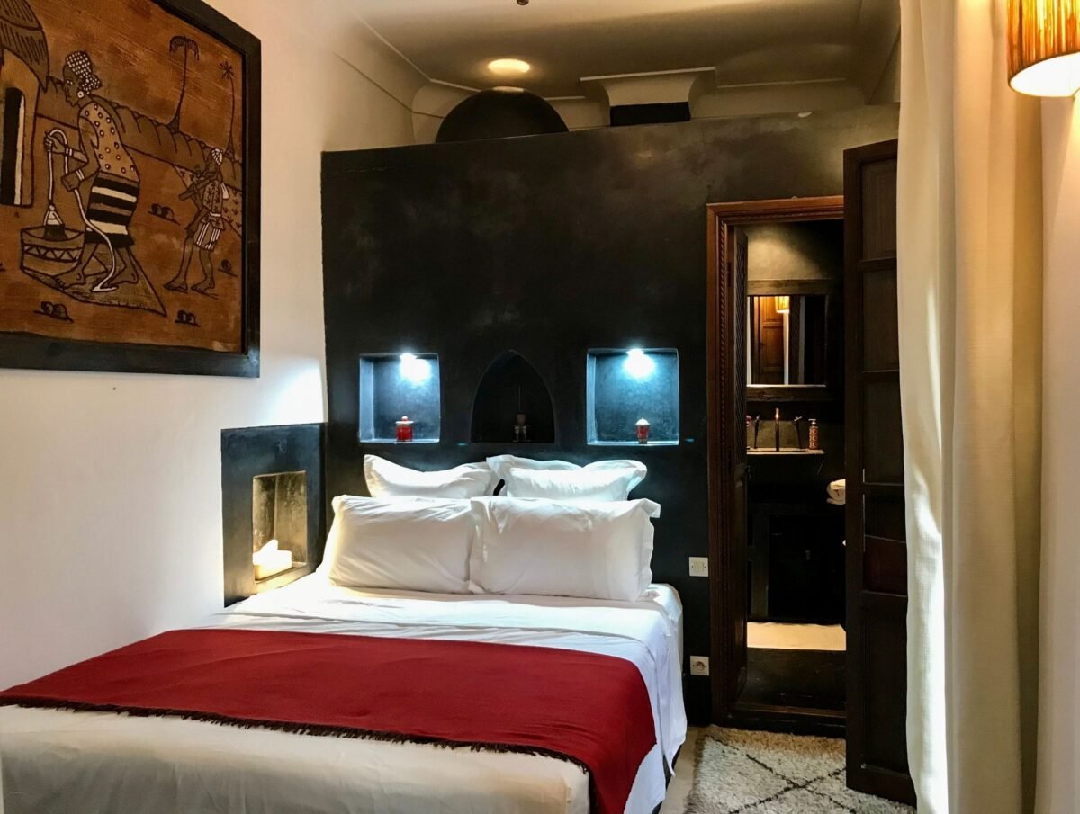 A cozy bedroom with a bed featuring white sheets, pillows, and a red throw blanket. There is a dark accent wall with built-in reading lights and small shelves. A painting hangs on the wall to the left, and a mirror reflects part of the room. A wooden door leads to a bathroom with a mirror, sink, and various toiletries. Curtains are partially visible on the right.