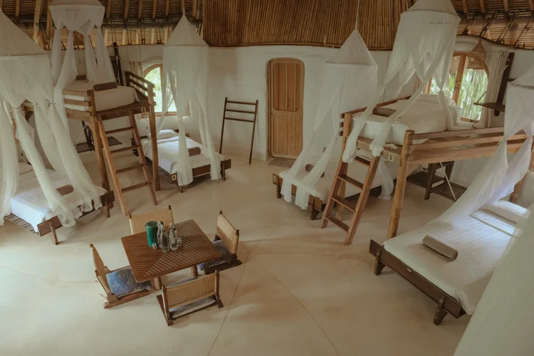 Interior of a rustic hostel with multiple wooden bunk beds adorned with white mosquito nets, arranged around a central wooden table with chairs, inside a bamboo and thatch structure.