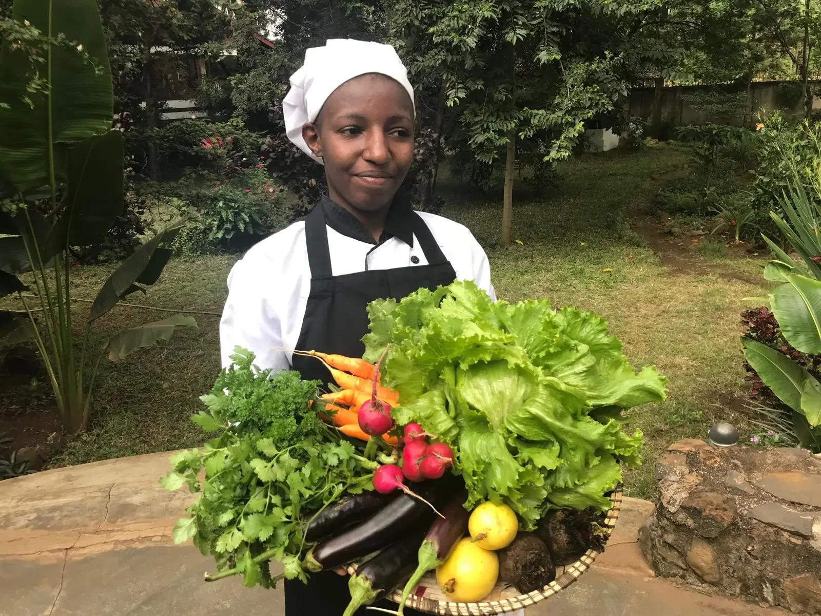 A young chef in a white uniform and black apron holding a basket of fresh vegetables outdoors, surrounded by greenery.