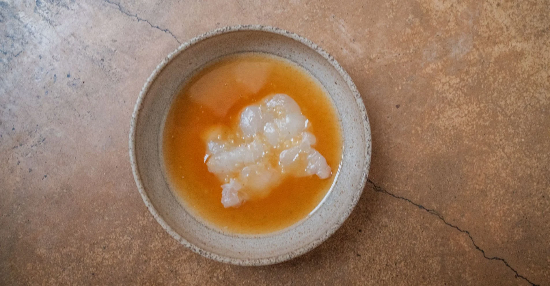 A ceramic bowl containing a mixture of clear liquid with white jelly-like chunks, placed on a brown, cracked surface.