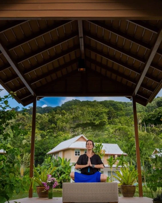 A woman practicing yoga or meditation on a porch with lush green hills in the background.