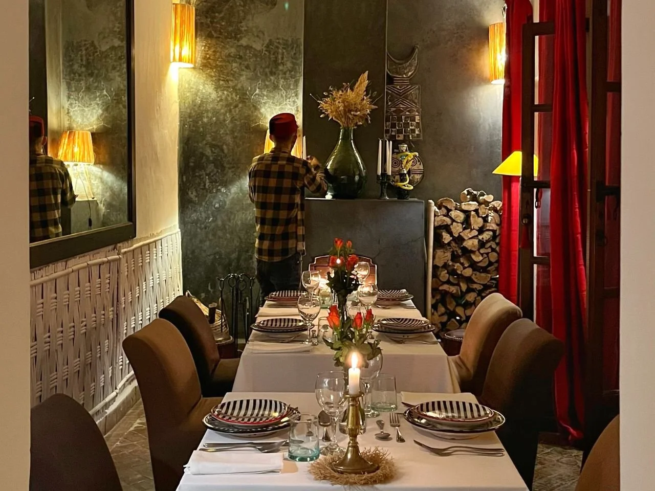 A dining room with a long table set for six, featuring plates with black and white stripes, glasses, and a floral centerpiece with orange flowers. In the background, a person with a red cap and checkered shirt stands near a fireplace decorated with a large green vase, candles, and a pile of firewood. The room has warm lighting, gold lamps on the walls, and red curtains.