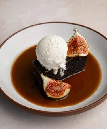 Chocolate cake topped with a scoop of vanilla ice cream, garnished with fresh fig slices and served with caramel sauce in a white bowl