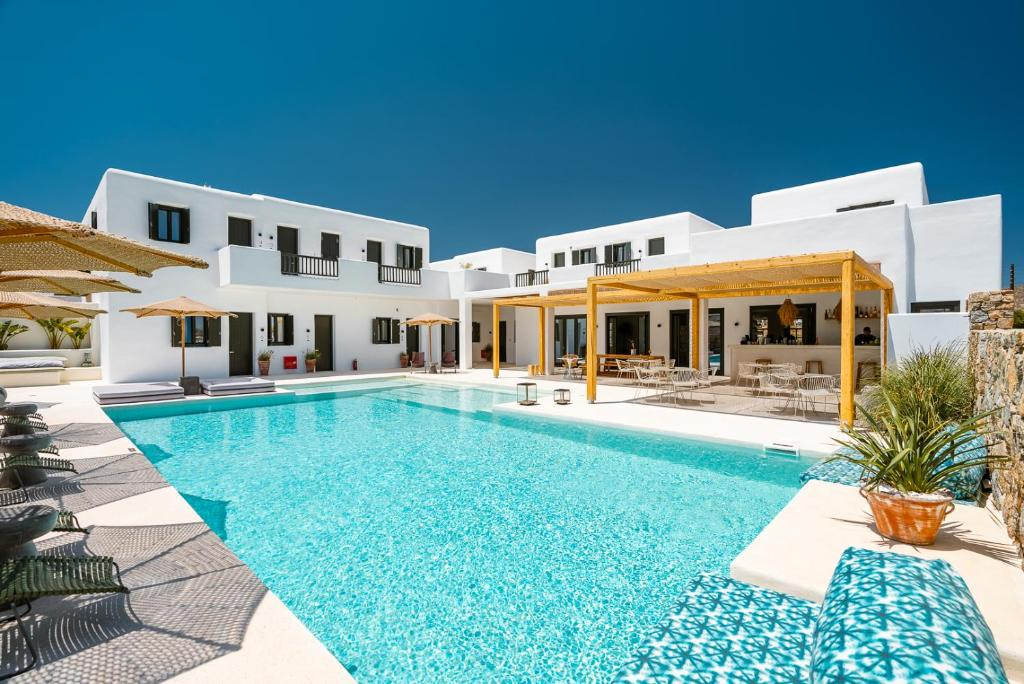 A luxurious white modern building with a swimming pool in front, surrounded by sun loungers and umbrellas under a clear blue sky.