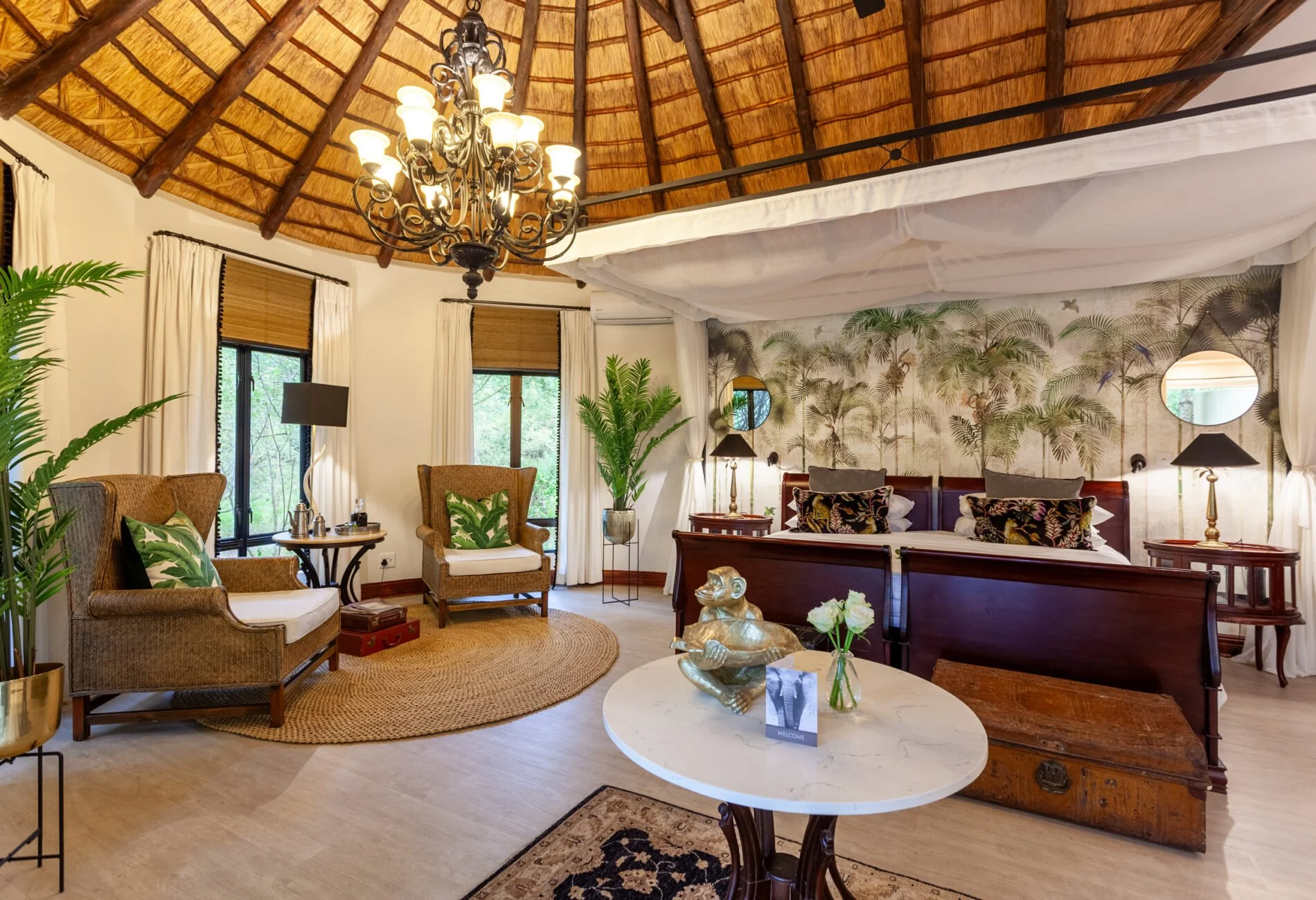 Interior of a tropical-style hotel room with a thatched roof ceiling, chandelier light fixture, large windows with white curtains, tropical wallpaper, and comfortable furniture including a bed with decorative pillows, armchairs, and small tables.
