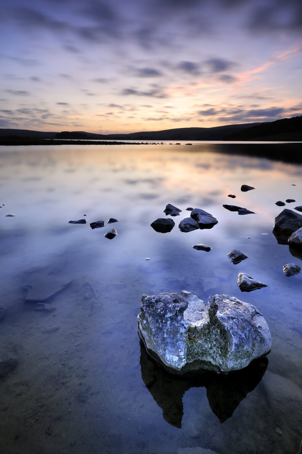 A peaceful lake at sunset with a sky filled with clouds. Rocks are scattered in the shallow water, with one large rock in the foreground reflecting in the calm water.