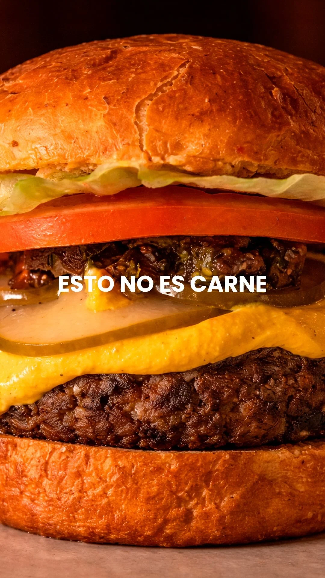 A tall burger with multiple layers of ingredients including lettuce, tomato, cheese, and meat, with the Spanish text 'ESTO NO ES CARNE' overlaid in the center.