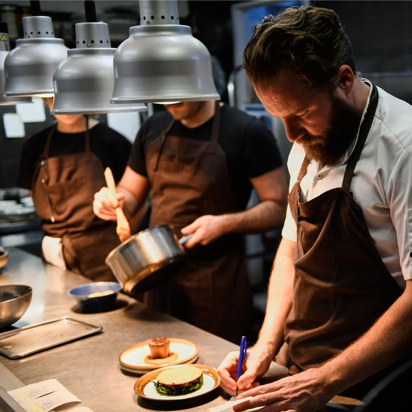Chefs in a professional kitchen working on plated dishes, including a sandwich with greens and a small dessert, under hanging lamps.