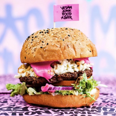 Vegan burger with lettuce, tomato, plant-based patty, pickles, and pink sauce on a bun, with a pink flag that reads 'Vegan Junk Food Bar'.