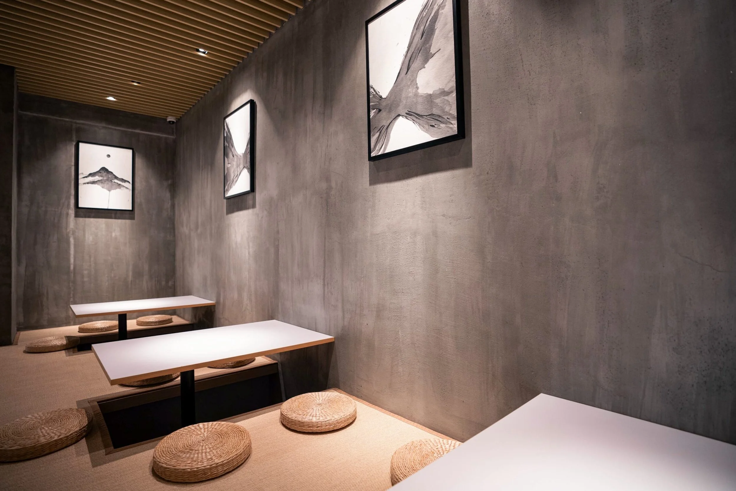 Interior of a modern room with a concrete wall, featuring three framed black and white landscape paintings. Low tables with woven round cushions arranged around them are on a beige carpet, with a wooden slatted ceiling with built-in lighting.