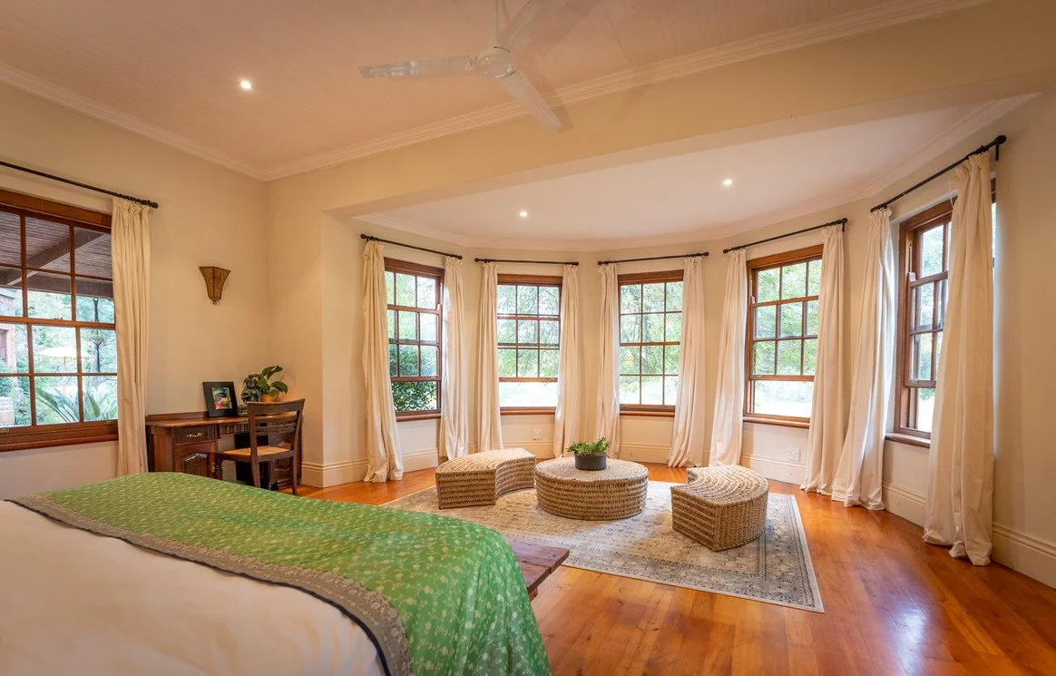 Bright bedroom with a bay window area, white curtains, wooden floors, a bed with green bedding, small wicker stools, a round wicker table with a plant, and a small wooden desk with a chair.