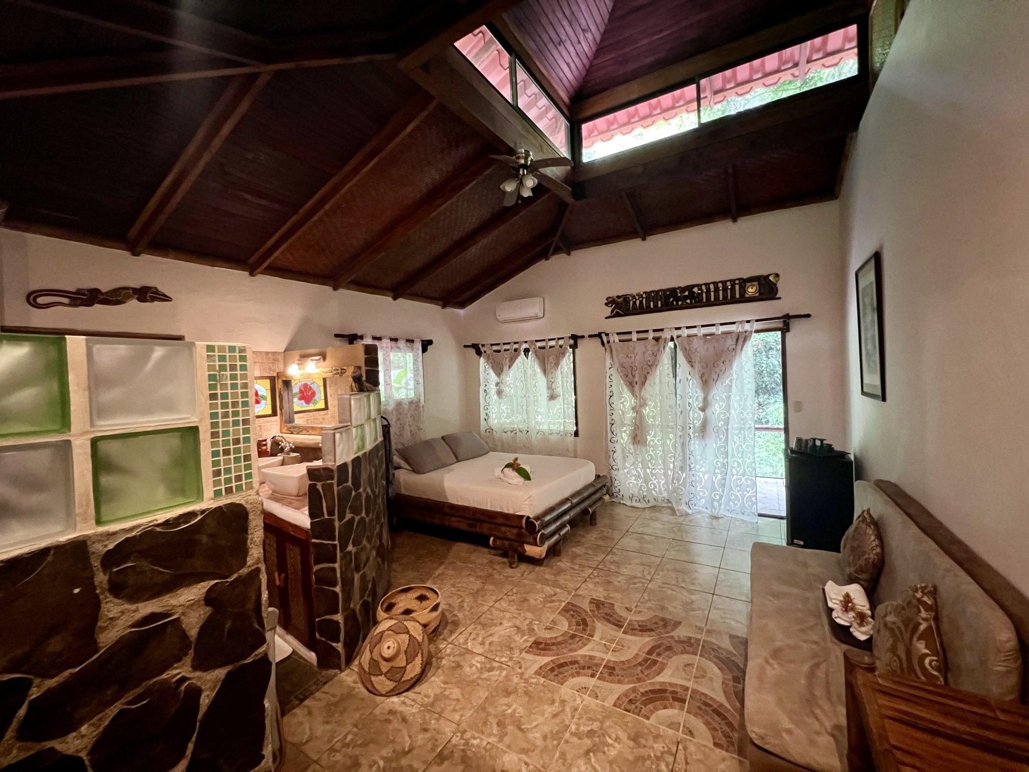 A cozy room with a vaulted wood ceiling and beige tiled floor, featuring a bed, a sofa, a small black refrigerator, and large windows with sheer curtains, revealing a balcony outside.