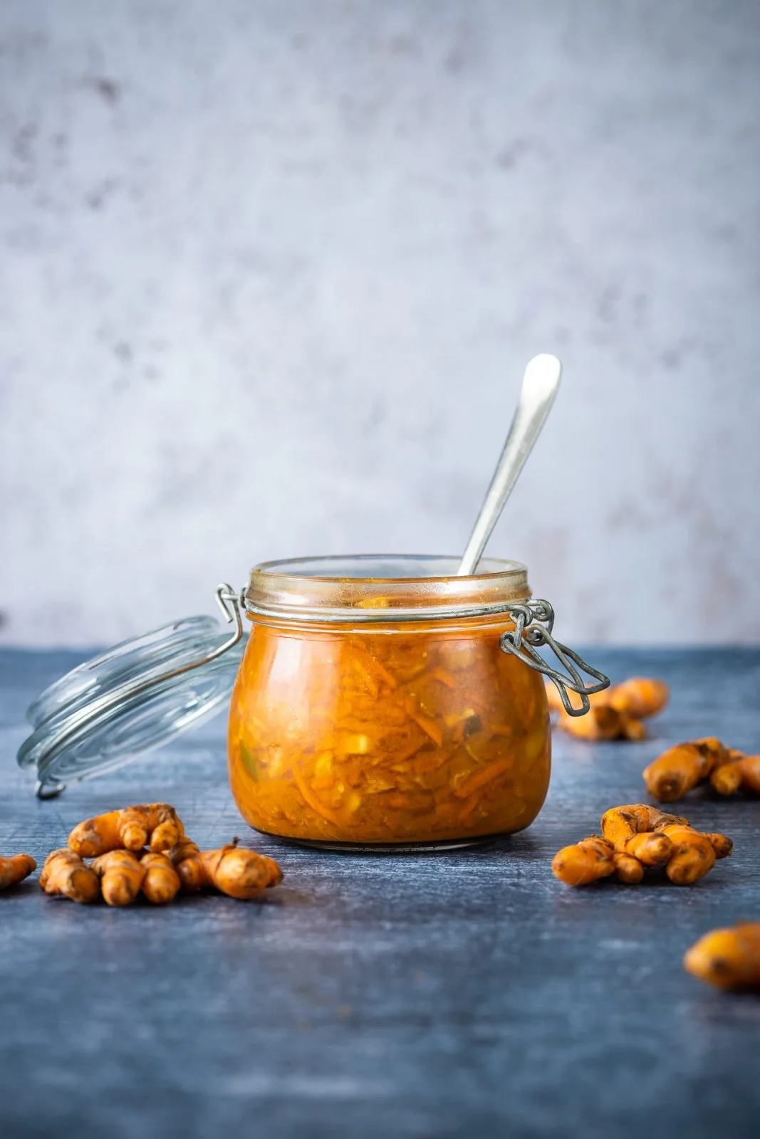A glass jar filled with turmeric pickle, surrounded by fresh turmeric roots on a dark wooden surface.