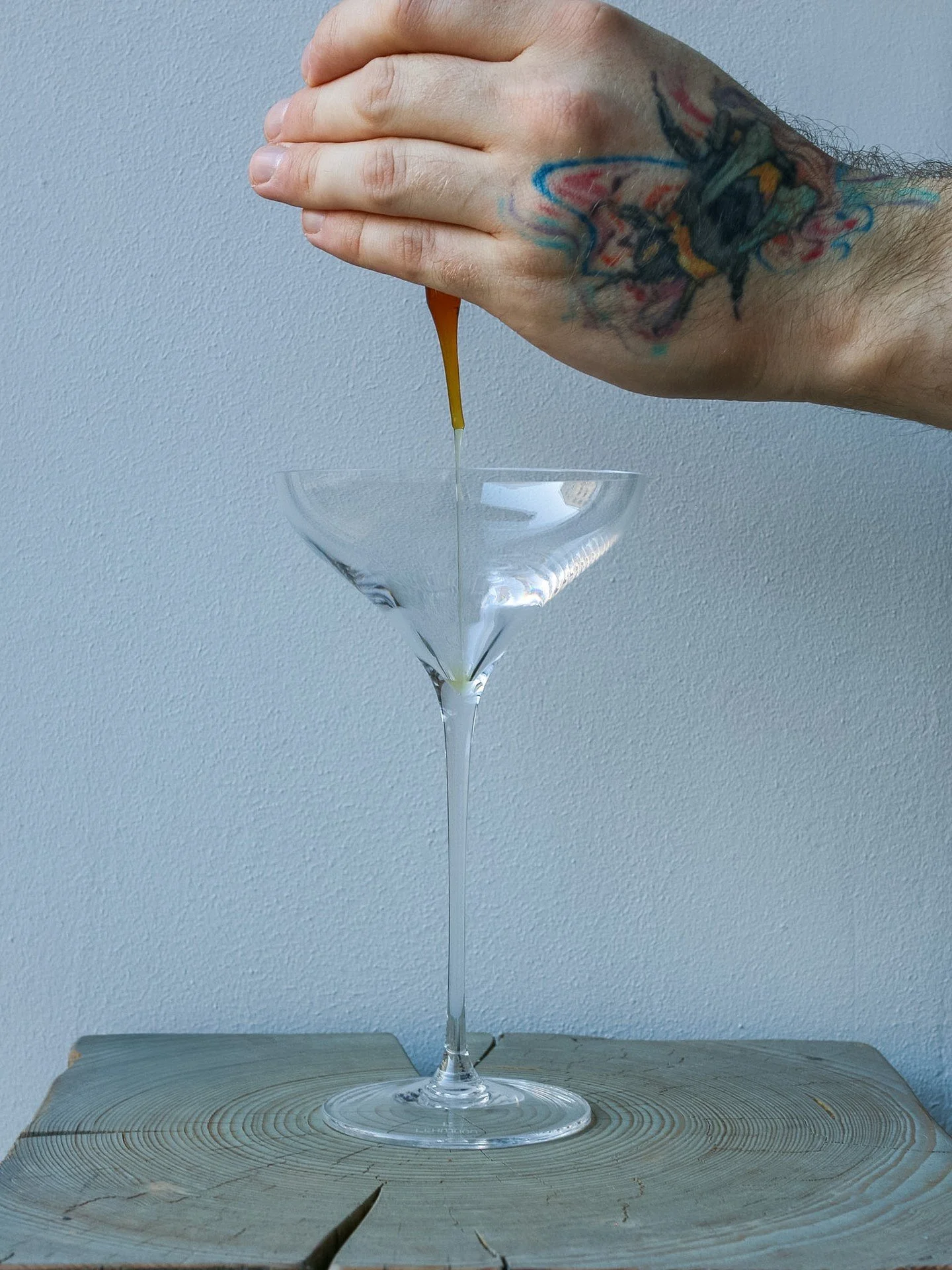 A hand with a colorful tattoo on the wrist holding a dropper and pouring a liquid into a martini glass on a wooden surface against a plain wall.