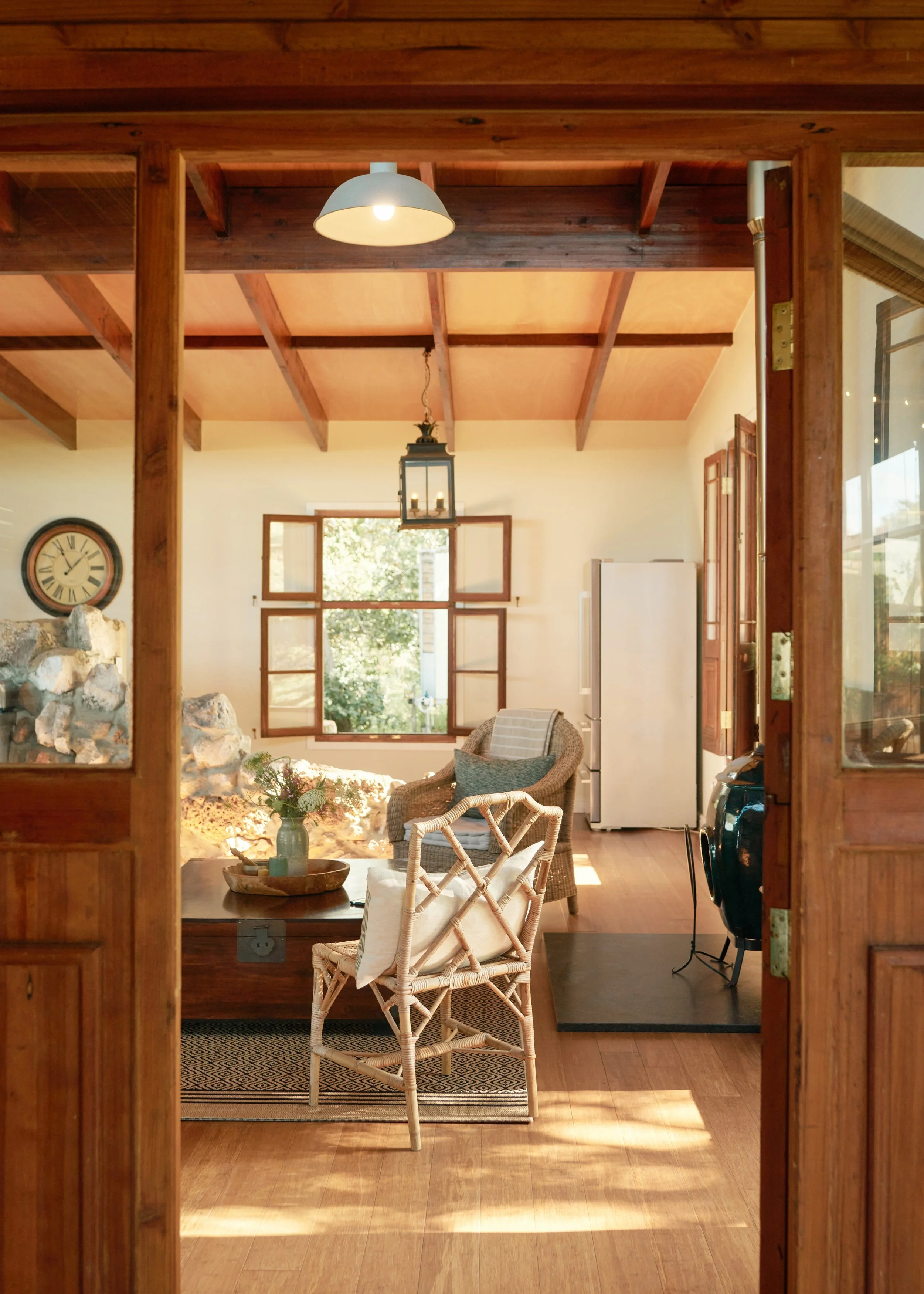 view of a cozy living room with wooden beams and soft lighting, featuring a dining table with a vase of flowers, a white chair, an open window, a clock, and kitchen appliances like a refrigerator, with natural sunlight streaming in.