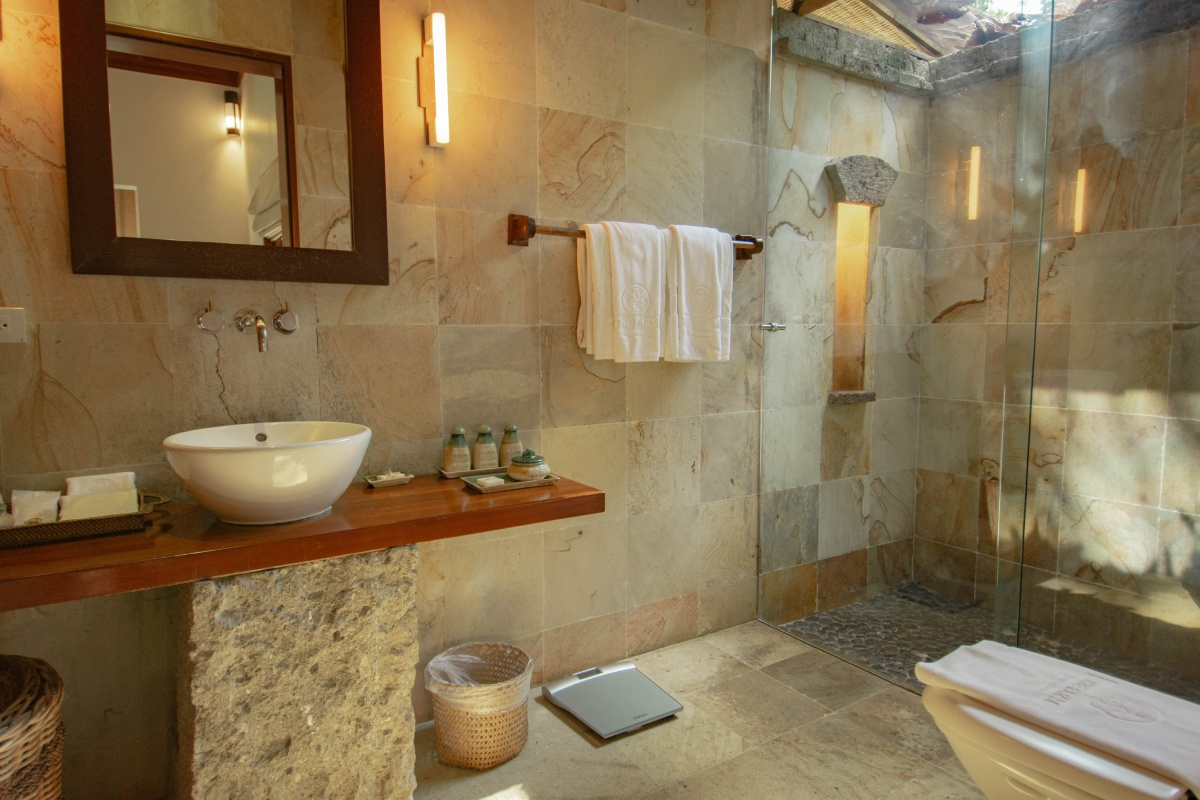 Bathroom with stone walls, a walk-in shower with glass door, a wooden countertop with a white vessel sink, a mirror, wall-mounted lights, a towel rack with white towels, a basket, a scale on the floor, and a rolled towel on the toilet.