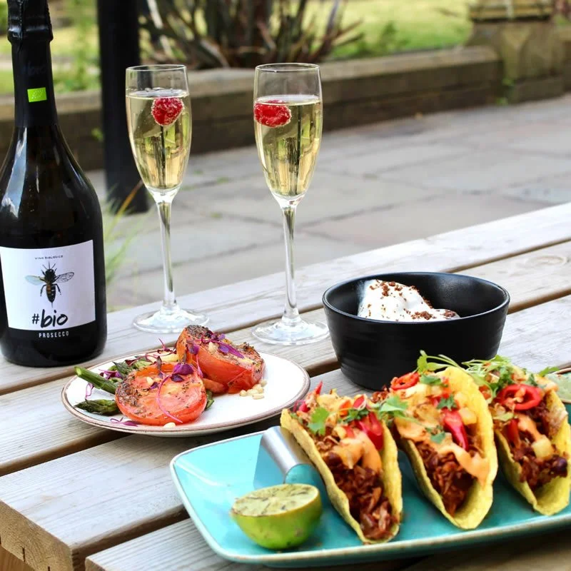 A picnic table with two glasses of champagne garnished with raspberries, a bottle of Prosecco, grilled stuffed tomatoes on a plate, three tacos with meat, cheese, and vegetables, a lime wedge, and a bowl of dessert topped with whipped cream.