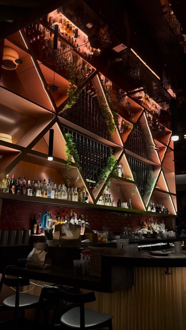 Modern bar interior with geometric shelves holding bottles, green plants, and decorative items, illuminated by warm lighting.