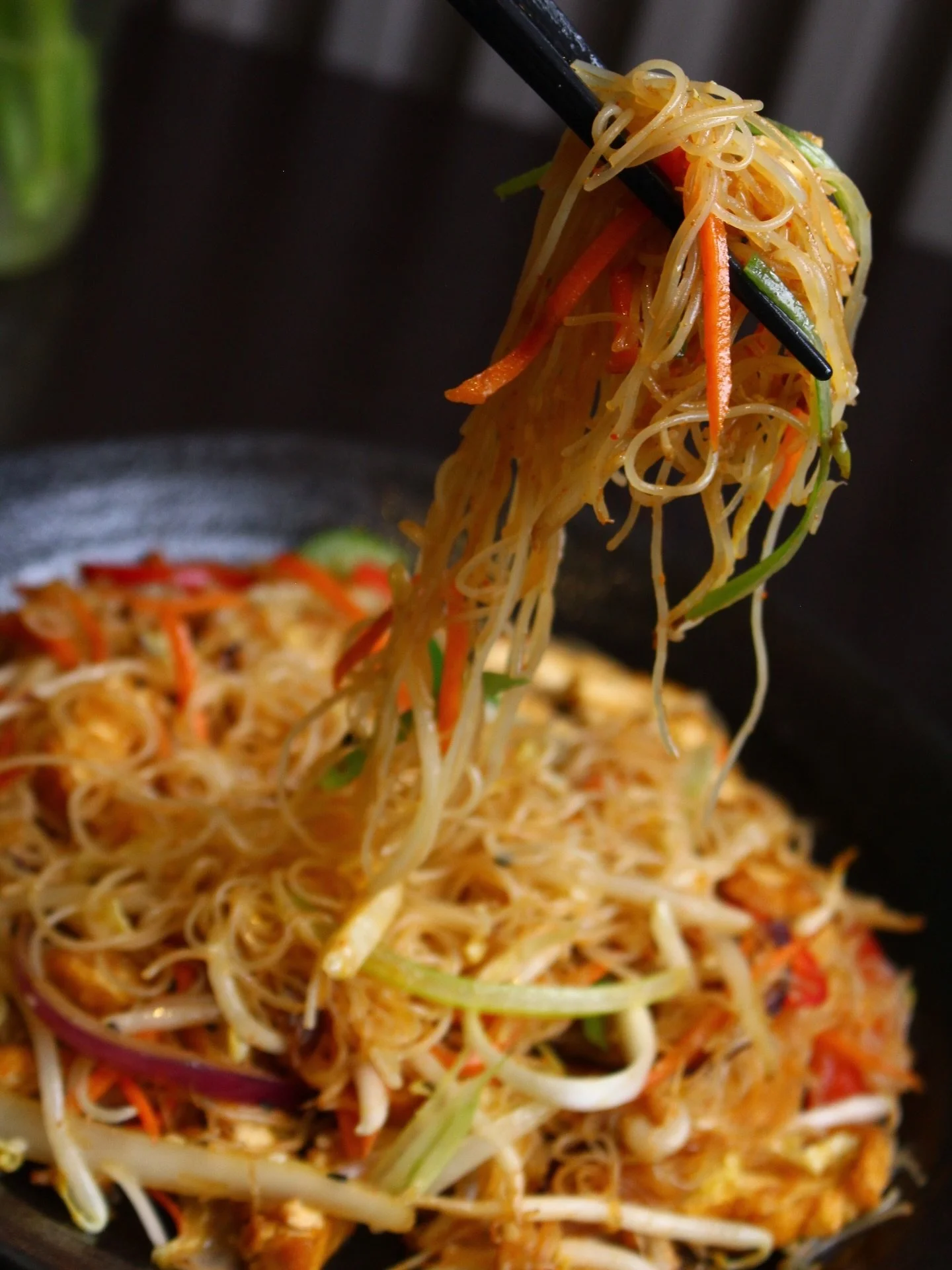 Close-up of stir-fried noodles with vegetables, including carrots, green onions, and bean sprouts, being lifted with chopsticks.