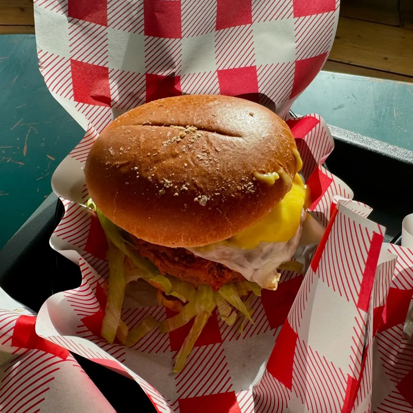A fried chicken sandwich with a golden bun, lettuce, fried chicken patty, melted cheese, and mayonnaise, served in red and white patterned paper.