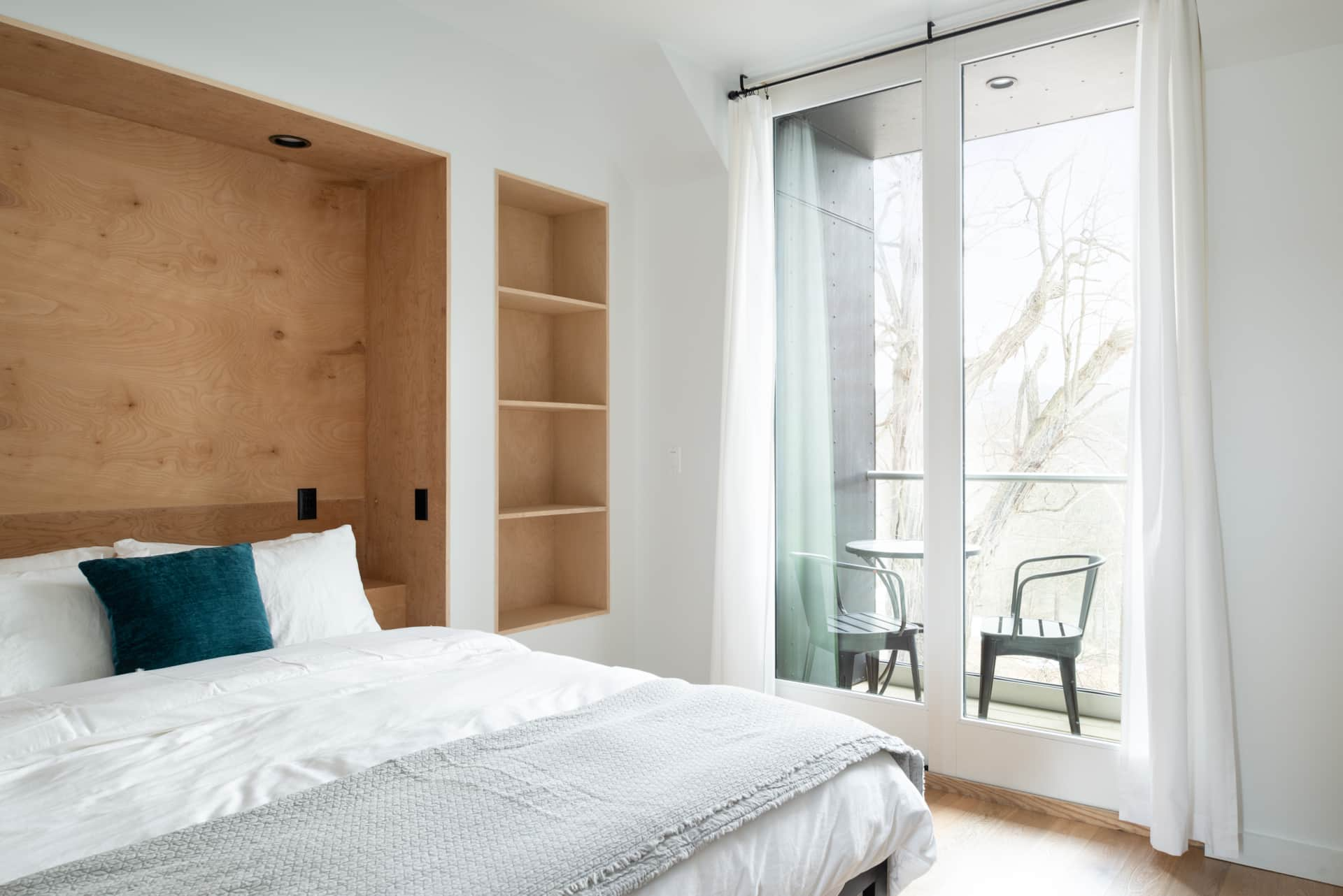 Bedroom with a bed, white bedding, teal pillow, built-in wooden headboard and shelves, large window with sheer curtains, balcony with two black chairs, trees outside, natural sunlight.