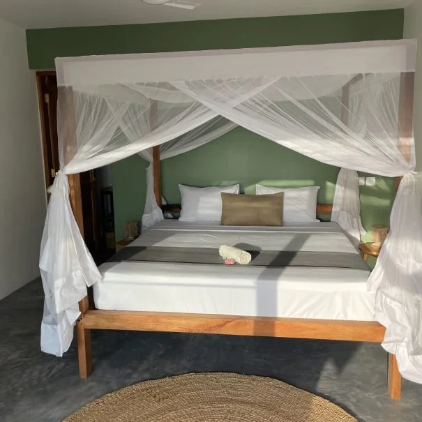 A four-poster bed with white mosquito netting in a room with green walls and gray flooring. The bed has white pillows, a beige throw pillow, and a folded towel and flower on top.